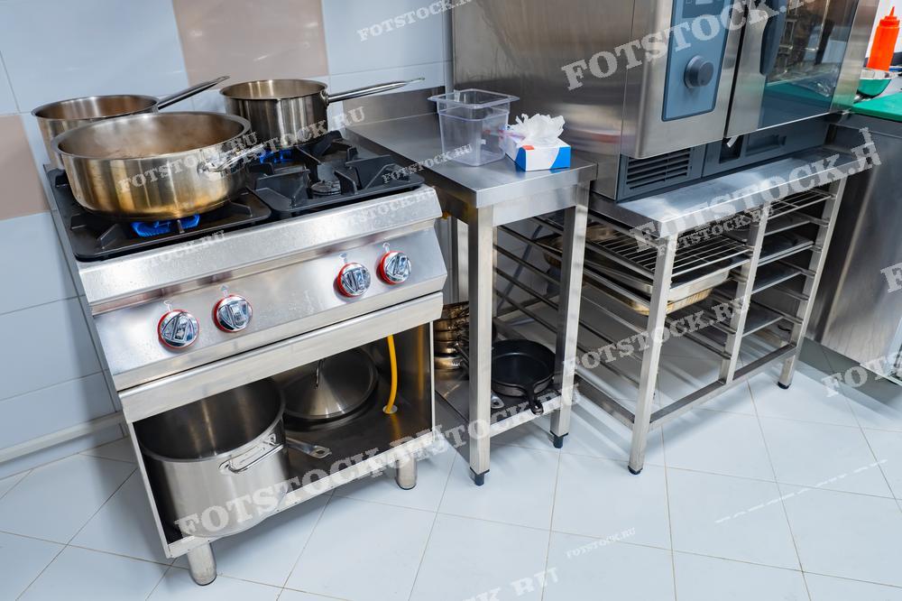 На изображении показана профессиональная кухня с плитой, на которой стоят кастрюли, и различным кухонным оборудованием. Стальные поверхности и функциональные элементы подчёркивают чистоту и организованность рабочей зоны. Фотография иллюстрирует процесс приготовления пищи в ресторане или кафе.